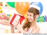 The image shows a young girl wearing a white crown and a pink dress, holding a red gift box with white polka dots. She is smi...