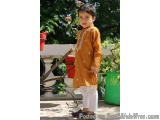 The image shows a young boy standing on a patio with a potted plant in the background. He is wearing a traditional Indian out...