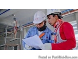 The image shows two men wearing hard hats and safety vests in a factory setting. They are both looking at a blueprint or docu...
