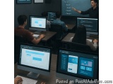 The image shows a group of people sitting at desks in front of multiple computer monitors. The desks are arranged in a semi-c...