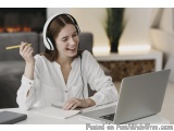The image shows a young woman sitting at a desk with a laptop and a notebook in front of her. She is wearing a white blouse a...