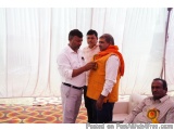 The image shows three men standing in a room with white curtains in the background. The man on the left is wearing a white sh...