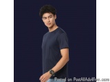 The image is a portrait of a young man standing against a dark blue background. He is wearing a navy blue t-shirt and light b...