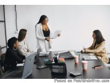 The image shows a group of four women sitting around a conference table in a meeting room. The woman in the center is standin...
