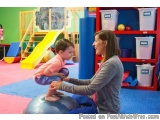 The image shows a young boy and a woman in a playroom. The boy is sitting on a blue exercise ball and is holding onto the bal...