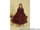The image shows a young girl standing in a room with a beige wall and a vase of flowers in the background. She is wearing a l...