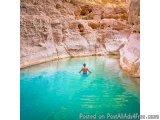 The image shows a man swimming in a beautiful turquoise-colored pool of water. The pool is surrounded by a rocky cliff on one...