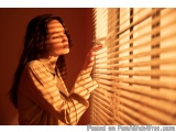 The image shows a young woman standing in front of a window with blinds. She is wearing a beige blouse and has long dark hair...