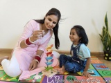 The image shows a young Indian mother and her young daughter sitting on a colorful rug in a room. The mother is wearing a pin...