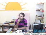 The image shows a young man sitting at a desk in an office. He is wearing a pink and black striped shirt and has a serious ex...