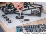 The image shows a person's hand holding a screwdriver and working on a gas stove top. The stove top has four burners and four...