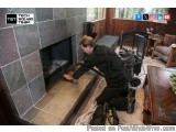 The image shows a man in a living room, kneeling on the floor in front of a fireplace. He is wearing a black shirt and pants...