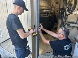 The image shows two men working on a metal door in a workshop. The man on the left is wearing a black t-shirt and a baseball...