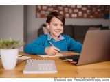 The image shows a young boy sitting at a wooden desk with a laptop in front of him. He is wearing a blue sweater and has a sm...