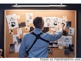 The image shows a man standing in front of a cork board with various photographs and postcards pinned to it. The man is weari...
