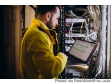 The image shows a man in a yellow jacket working on a laptop in a server room. He is standing in front of a rack of servers a...
