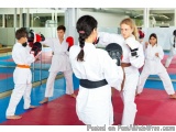 The image shows a group of children in a martial arts class. They are wearing white karate uniforms and red and black belts....