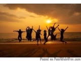 The image shows a group of people on a beach at sunset. The sky is orange and yellow, with the sun setting in the background....