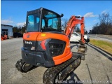The image shows an orange and black Kubota mini excavator parked on the side of a road. The excavator has a bucket attached t...