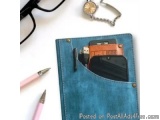 The image shows a blue denim notebook with a pocket on the left side. Inside the pocket, there is a black pen and a small ora...