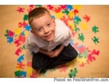 The image is a portrait of a young boy sitting on the floor covered in colorful puzzle pieces. He is wearing a white shirt an...
