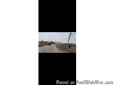 The image is a photograph of a road with a truck driving on it. The road appears to be in a rural area with trees and bushes...