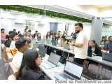 The image shows a group of people sitting in a lecture hall with laptops on their laps. The room has a high ceiling with gree...