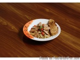 The image shows a plate of food on a wooden table. The plate is white with an orange rim and is filled with a dish of noodles...