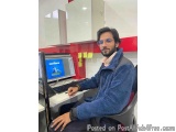 The image shows a young man sitting at a desk in front of a computer. He is wearing a blue jacket and has a beard and glasses...