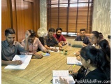 The image shows a group of nine people sitting around a wooden table in a meeting room. They are all engaged in a discussion...