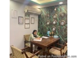 The image shows a woman sitting at a wooden desk in an office. She is wearing a white blouse and appears to be working on a p...