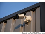 The image shows a security camera mounted on the side of a building. The camera is white in color and has a round lens. It is...