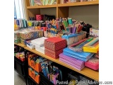 The image shows a large collection of colorful stationery items on a wooden shelf in a classroom. The shelf is filled with a...