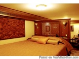 The image shows a bedroom with a large bed in the center. The bed is covered with a beige comforter and several orange pillow...