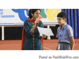 The image shows a young boy and a young woman standing on a stage. The woman is holding a microphone and appears to be giving...