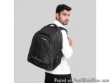 The image shows a young man wearing a black backpack with a red logo on the front. He is standing with his left hand on his h...