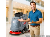 The image shows a young man in a blue polo shirt and khaki pants standing in a shopping mall. He is holding a floor scrubber...