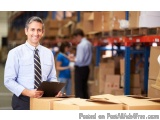 The image shows a man in a warehouse, holding a clipboard and smiling at the camera. He is wearing a blue shirt and tie and i...