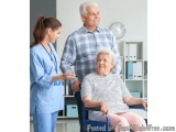 The image shows an elderly woman in a wheelchair being assisted by a nurse. The nurse is wearing a blue uniform and is standi...