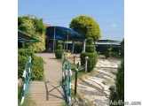 The image shows a wooden walkway with a green railing on the right side. The walkway is surrounded by a garden with shrubs an...