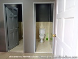 The image shows a small bathroom with a white toilet and a gray door. The walls are painted in a light grey color and the flo...