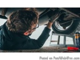 The image shows a man working on a car under the underside of a vehicle. He is wearing a black jacket and glasses and is hold...