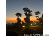 The image is a photograph of a beautiful sunset over a hillside. The sky is a mix of orange, yellow, and blue hues, with the...