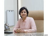 The image is a portrait of a woman sitting at a desk in an office. She is wearing a pink striped blouse and has short dark ha...