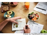 The image shows a person's hand holding a pen and writing on a diet plan book. The book is open and the person is sitting at...