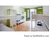 The image shows a room with multiple bunk beds arranged in a row. The bunk beds are white and have blue blankets and pillows...