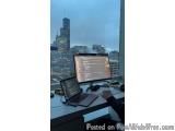 The image shows a desk with two laptops and a desktop computer on it. The desk is in front of a large window with a view of a...