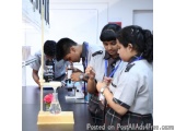The image shows a group of four young students in a laboratory setting. They are wearing school uniforms and are gathered aro...