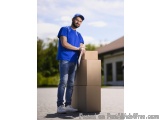 The image shows a young man standing in front of a house with a stack of cardboard boxes. He is wearing a blue t-shirt, blue...