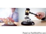 The image shows a hand holding a gavel over a miniature model of a house on a wooden table. The gavel is black and gold in co...